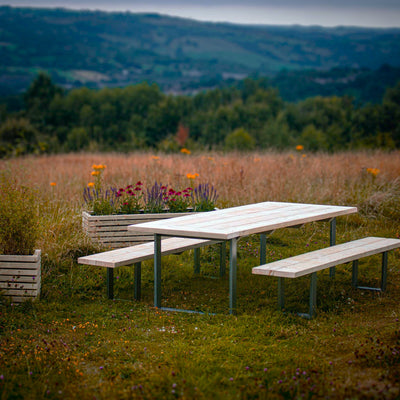 A Rustic Wooden Garden Table with Steel Box Legs and matching benches. Two wooden planters are set near the table set. The set is surrounded by a scenic landscape with rolling hills and a blue sky.