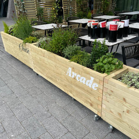 Wooden branded planters for Chase Arcade, a bar in Oxford Circus. The planters are filled with green plants and surround an outside dining area.