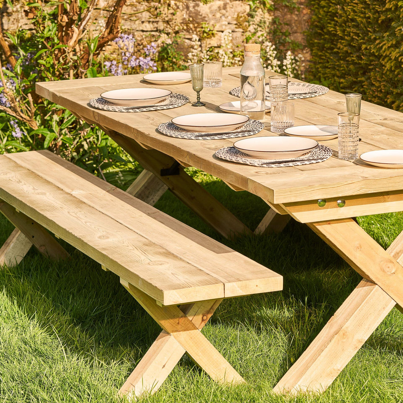 This chunky wooden rustic garden table is handcrafted from FSC-certified sustainable timber and features a bold 45mm thick tabletop with classic breadboard ends, complemented by sturdy X-style legs for a timeless farmhouse look. It's placed on a garden lawn and set with plates, cutlery and glasses.