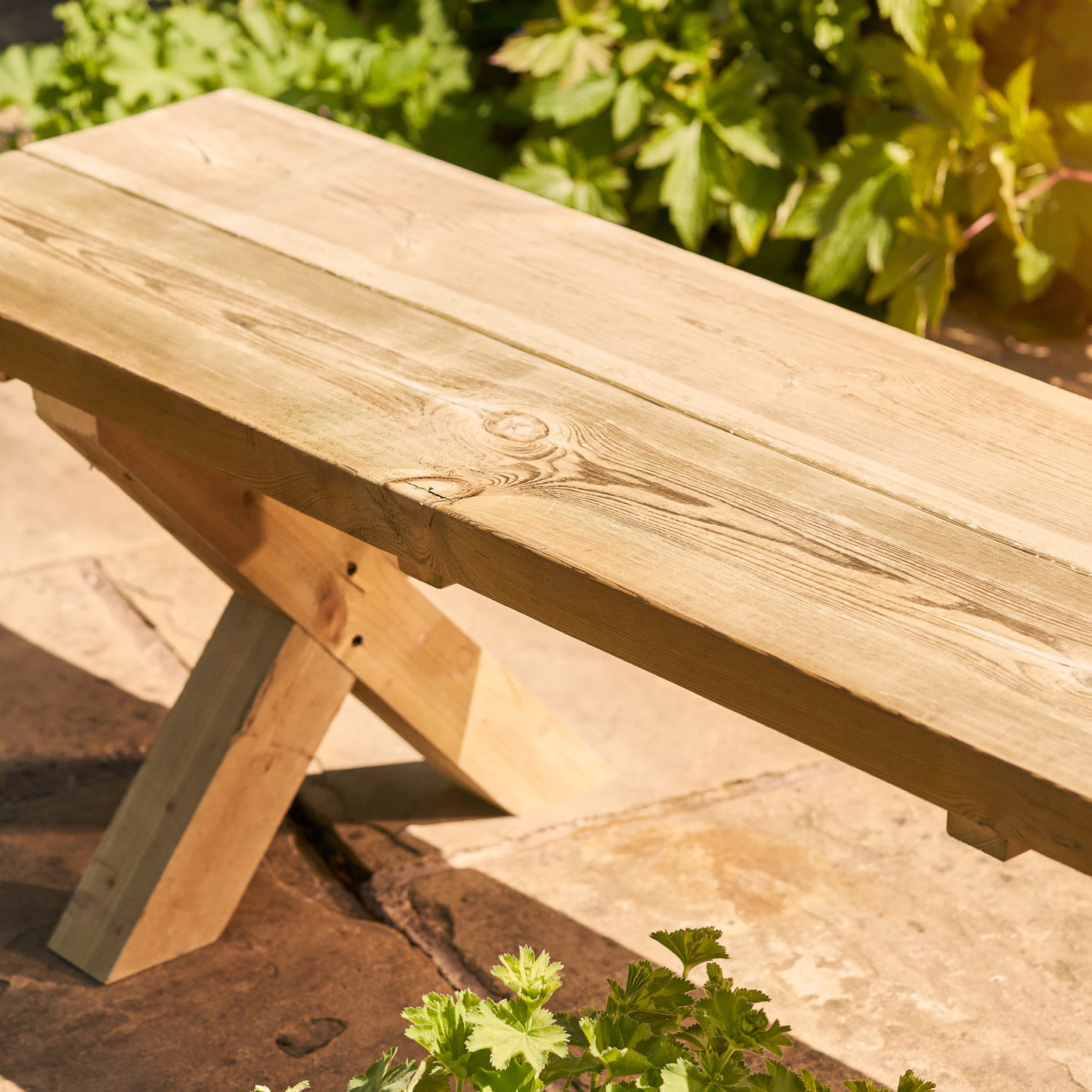 A close-up view of a wooden bench. This chunky rustic farmhouse style garden bench is handcrafted from FSC-certified sustainable timber, features a bold 45mm thick top and x shaped legs.