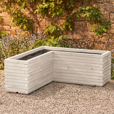 A wooden corner planter in a soft white stain featuring an L-shaped design that fits perfectly into any space. Positioned in front of a rustic stone wall with green climbing branches, this planter is crafted from durable FSC-certified timber and comes pre-lined and ready for planting.