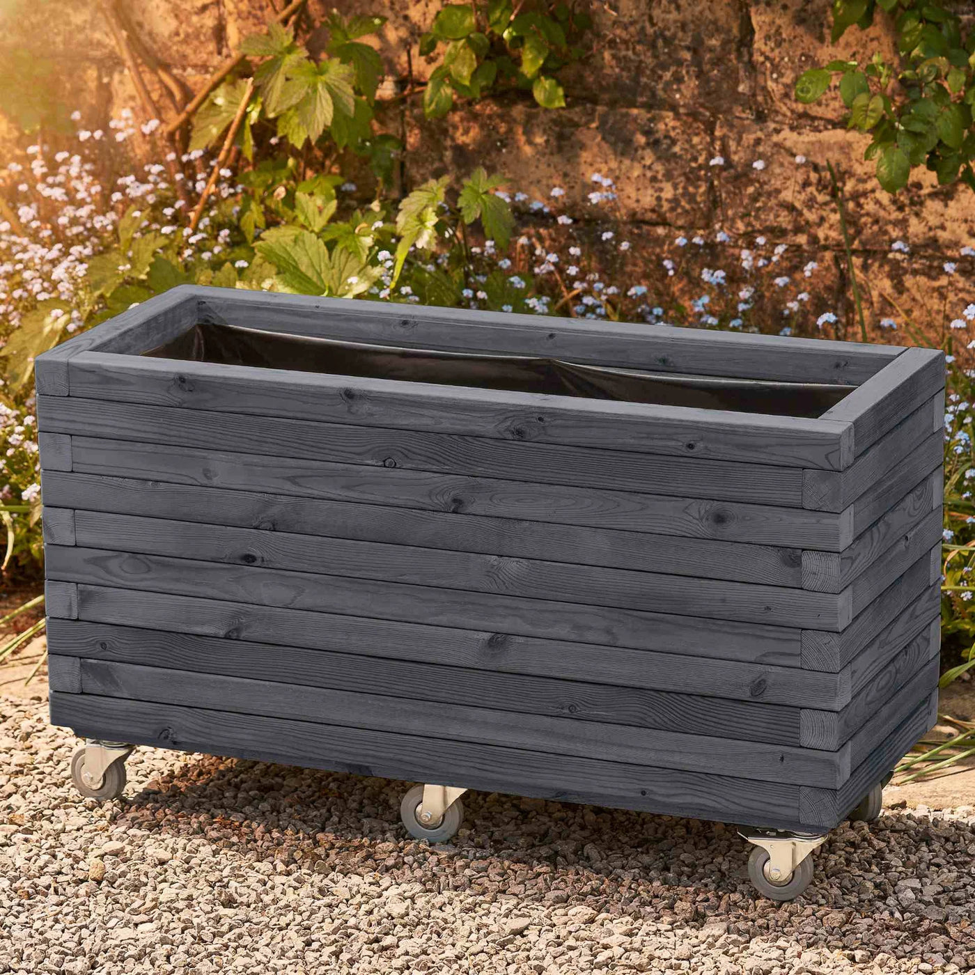 Grey Wooden Garden Planter with Wheels on gravel garden with wall behind and flowers.