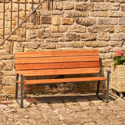 This hardwood garden bench, crafted from premium Sapele hardwood, offers exceptional durability and modern style. It sits in front of a stone wall by a wooden planter.