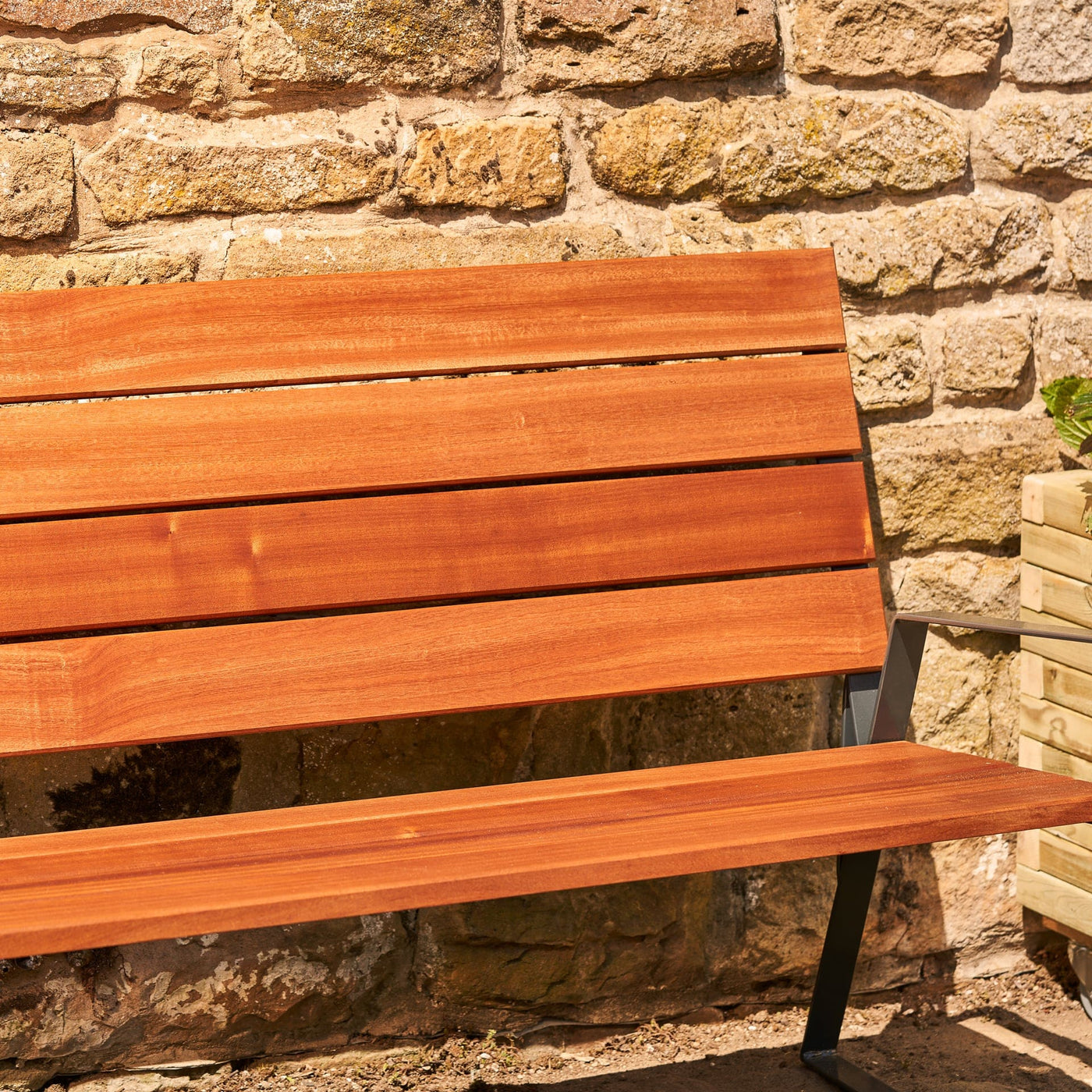 A close-up view of a premium outdoor garden bench made from premium sapele hardwood and robust UK steel and placed in front of a stone wall.