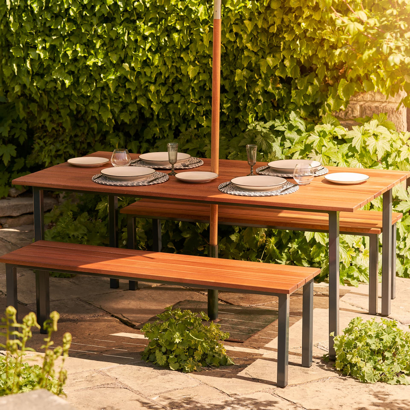 Hardwood Garden Table & Benches with parasol. The table is set with white plates, cutlery and glasses and sits on a patio in front of lush greenery.