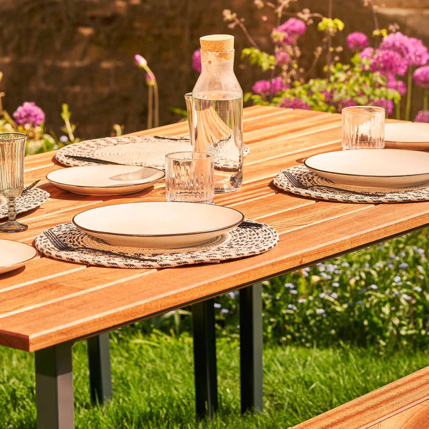 Hardwood Garden Table & Benches with parasol. The table is set with white plates, cutlery and glasses and sits on lush grass in front of a stone wall.