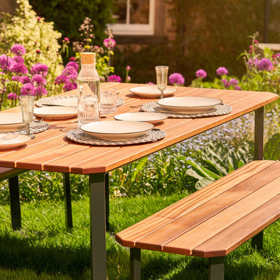 A corner close-up view of a Hardwood Outdoor Dining Set with an angled design and parasol. The table is set with plates, cutlery and glasses and sits on a lush garden lawn.