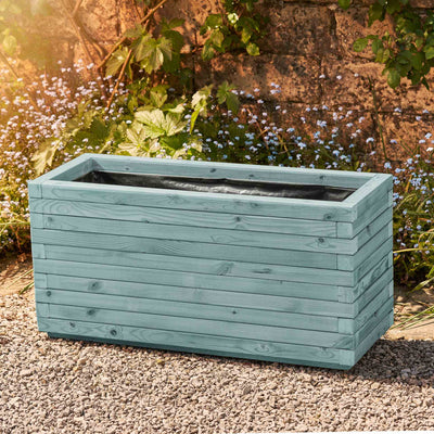Large rectangular garden planter in a beach hut blue finish, lined for added functionality. This durable wooden planter features a modern, sleek design and is placed against a wall with climbing green plants.
