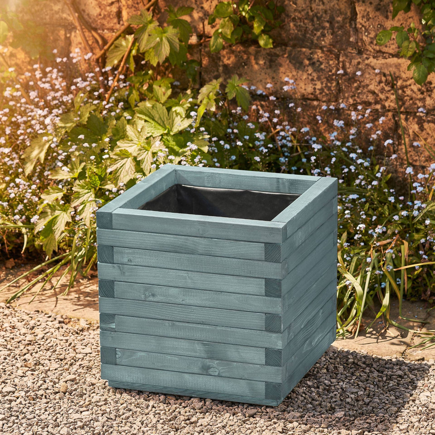 Large square wooden planter in a beach hut blue finish with a modern, sleek design. This durable planter is lined and ready for planting. Positioned against a wall with climbing green plants.