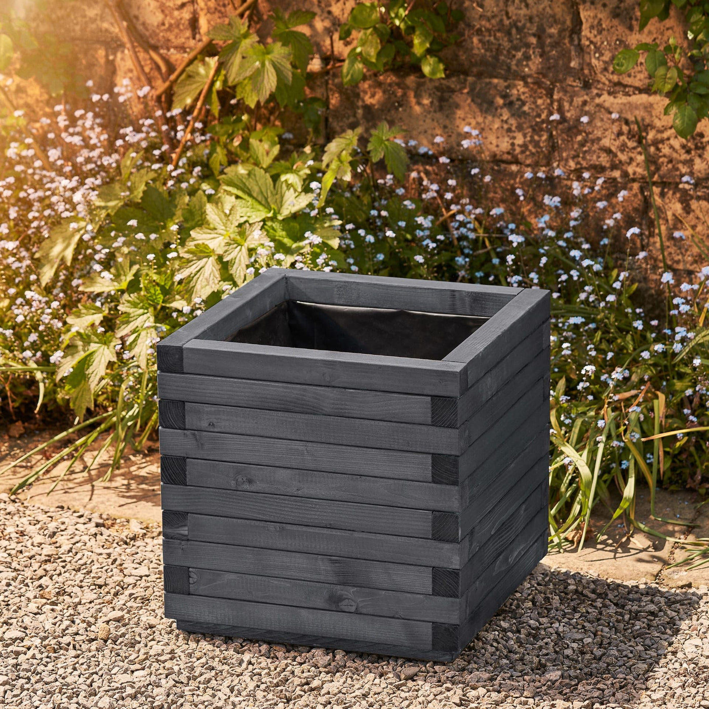 Large square wooden planter in an ironstone grey finish with a modern, sleek design. This durable planter is lined and ready for planting. Positioned against a wall with climbing green plants.