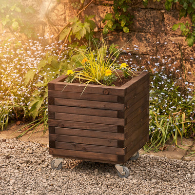 Large square wooden planter in a rich medium oak finish with a modern, sleek design. This durable planter is filled with vibrant yellow flowers and greenery, making it a striking addition to any garden. Positioned against a wall with climbing green plants.