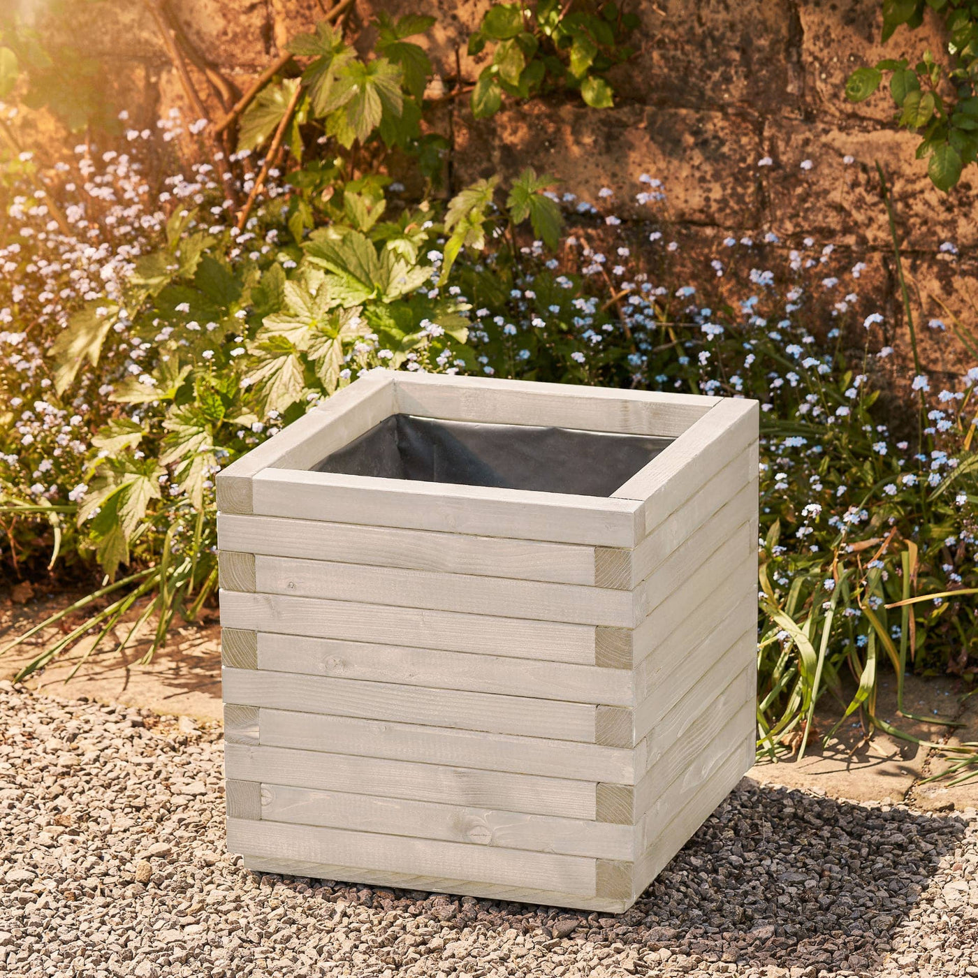 Large square wooden planter in a soft white finish with a modern, sleek design. This durable planter is lined and ready for planting. Positioned against a wall with climbing green plants.