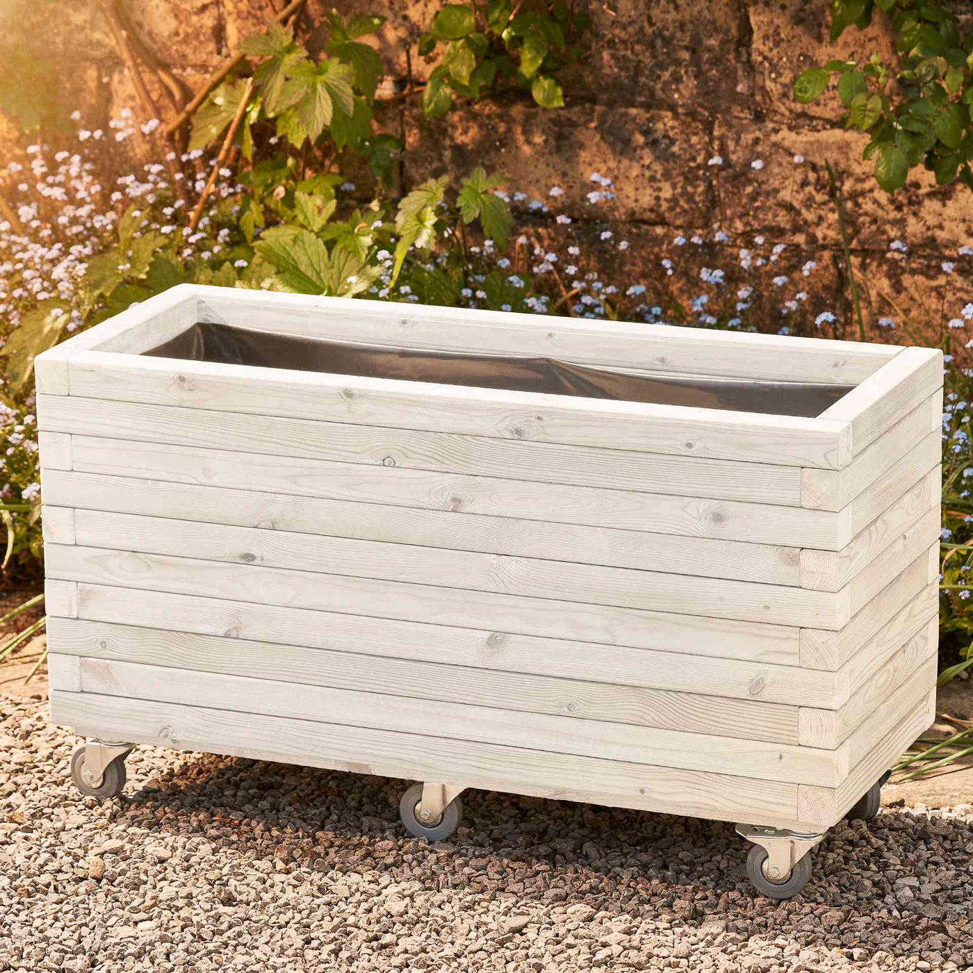 Large rectangular garden planter in a soft white finish, lined and fitted with wheels for added functionality. This durable wooden planter features a modern, sleek design and is placed against a wall with climbing green plants.