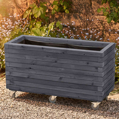 Large rectangular garden planter in an ironstone grey finish, lined and fitted with wheels for added functionality. This durable wooden planter features a modern, sleek design and is placed against a wall with climbing green plants.