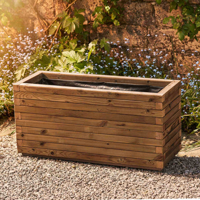 Large trough style garden planter in a medium oak finish, lined for added functionality. This durable wooden planter features a modern, sleek design and is placed against a wall with climbing green plants.