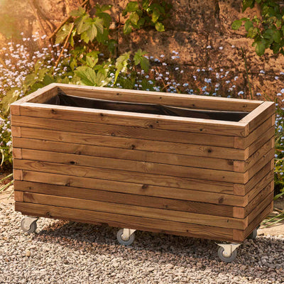 Large trough style garden planter in a medium oak finish, lined and fitted with castors/wheels for added functionality. This durable wooden planter features a modern, sleek design and is placed against a wall with climbing green plants.