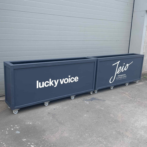 2 Grey wooden planters with commercial bar branding painted on the front in white. Lucky Voice and Jeio Prosecco. Commercial planters have durable wheels. Planters are empty and placed infront of grey shutters.