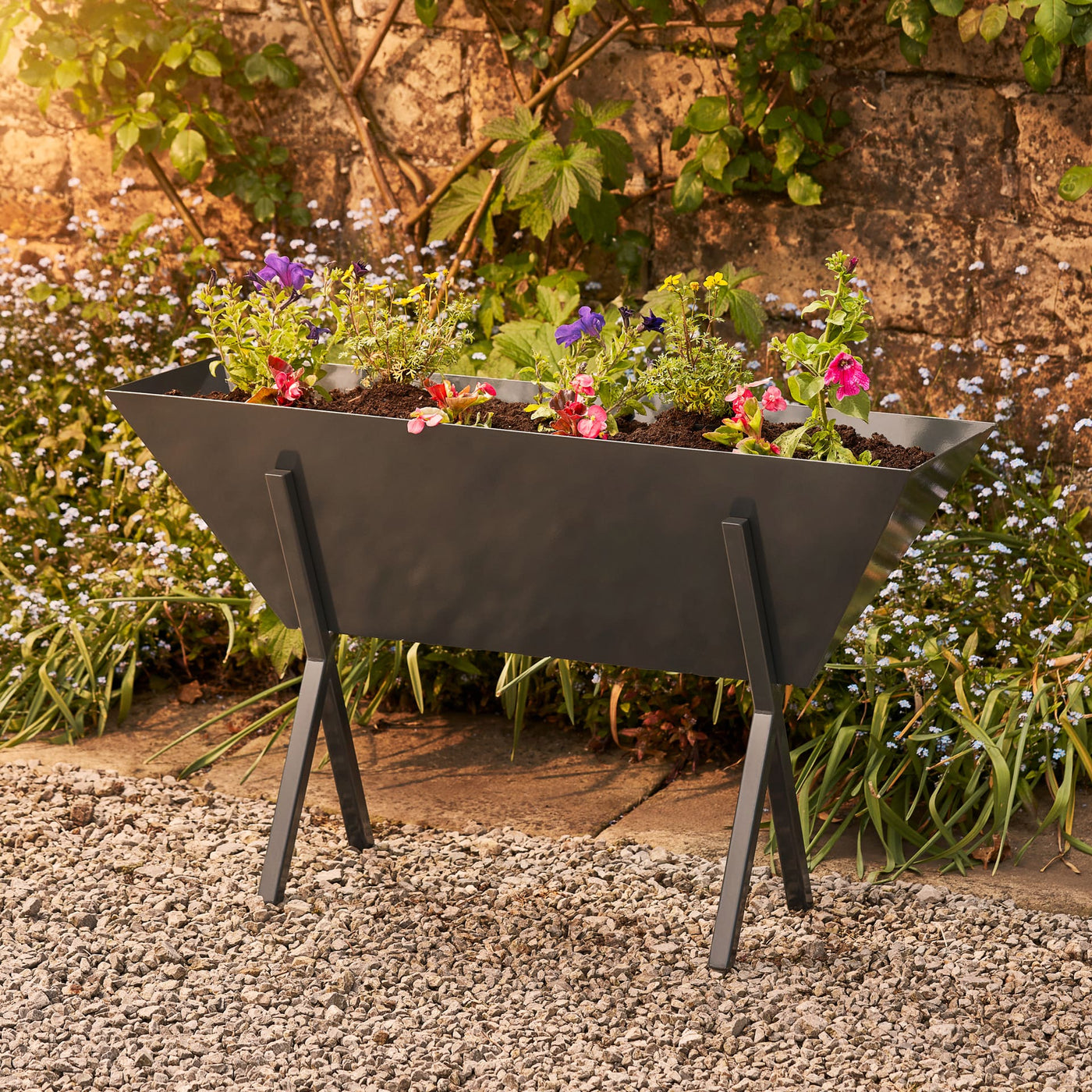 Metal trough planter in grey. UK forged fabricated steel coated in a resin epoxy coating for maximum durability. Placed in front of a stone wall and filled with bright pink and purple flowers.