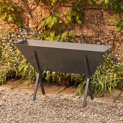 Metal trough planter in grey. UK forged fabricated steel coated in a resin epoxy coating for maximum durability. Placed in front of a stone wall.