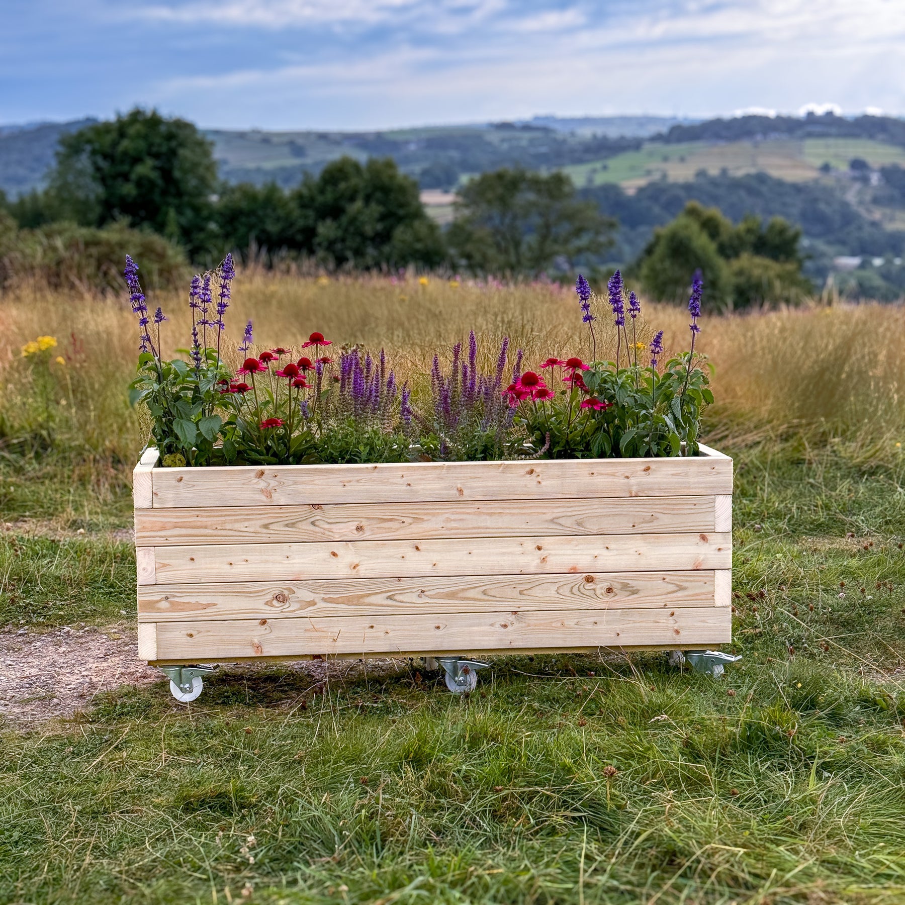 A rectangular wooden planter with casters, filled with vibrant flowers like lavender and red dianthus. The planter is set against a scenic backdrop of rolling hills in Matlock, Derbyshire.