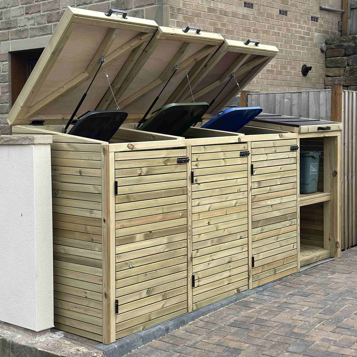 Wooden outdoor storage unit with hinged lids for trash cans against a brick wall.