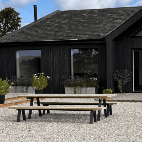 rustic farmhouse table on gravel garden in front of a black-clad house surrounded by plants with a blue sky in the background