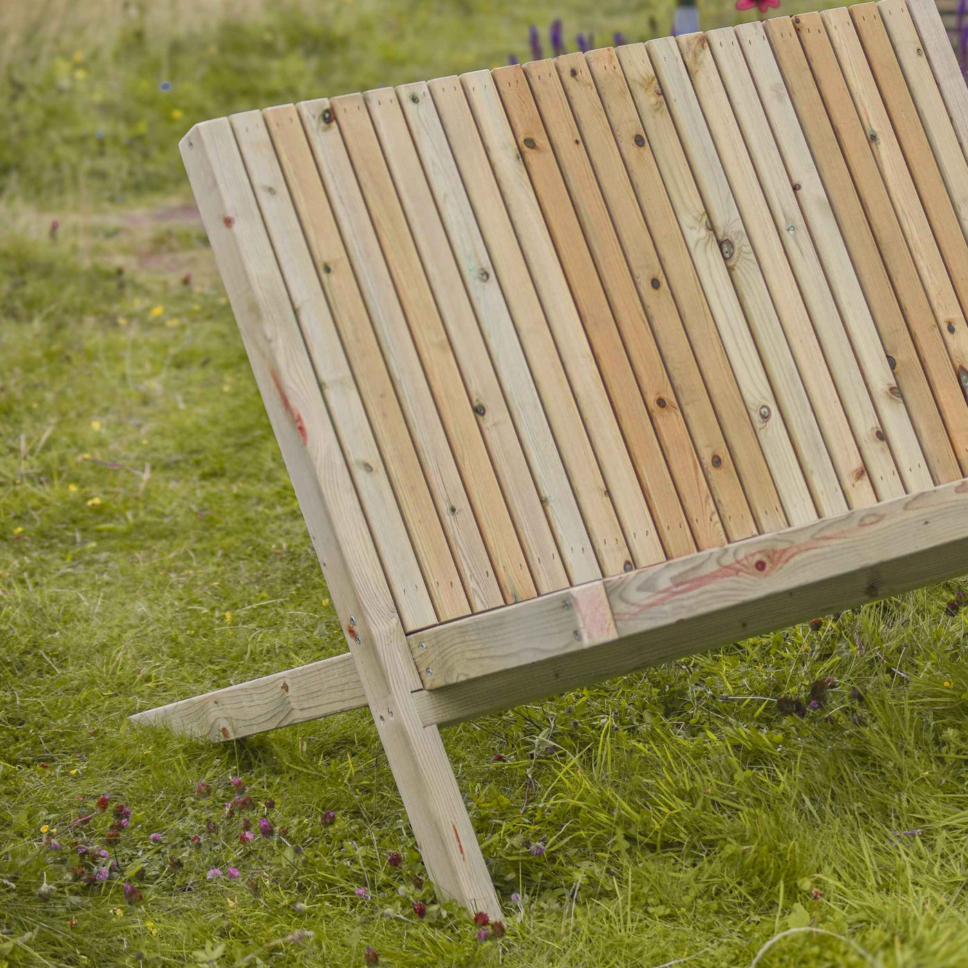 A close-up view of the back timber slats of a Rustic Garden Bench placed in a lush green garden. Made from durable fsc-timber. The bench is designed in an Adirondack style for comfort. 