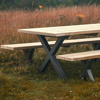 A rustic outdoor dining set featuring a long wooden table with metal X legs and two matching benches. The set is surrounded by lush greenery and a scenic landscape, making it perfect for entertaining guests or enjoying meals al fresco.