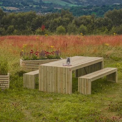 A durable, rustic wooden garden table set, perfect for outdoor dining and entertaining. The set features a long rectangular table with matching benches, all made from high-quality timber. The table and benches have a slatted design and are set with glasses and a bottle. Next to the table are two wooden planters.