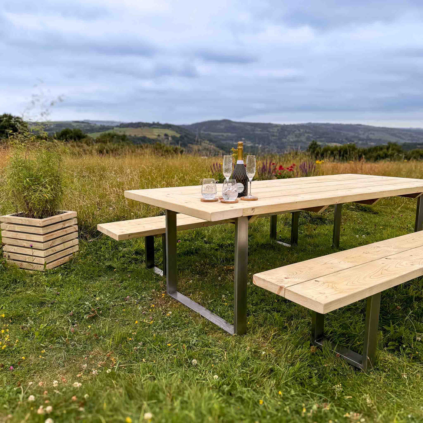 A Rustic Wooden Garden Table with Steel Box Legs and matching benches. A small wooden planter is placed nearby, adding a touch of greenery. The set is surrounded by a scenic landscape with rolling hills and a blue sky.