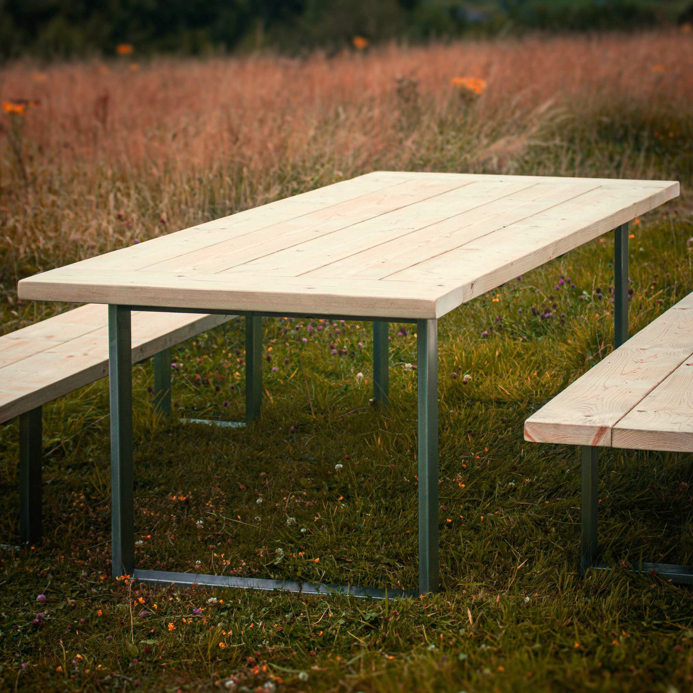 A close-up view of a Rustic Wooden Garden Table with Steel Box Legs and matching benches. The set is surrounded by a scenic landscape of long lush grass.