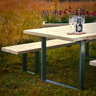 A Rustic Wooden Garden Table with Steel Box Legs and matching benches. A small wooden planter is placed nearby, adding a touch of greenery. The table is set with four iced glasses and a bottle.