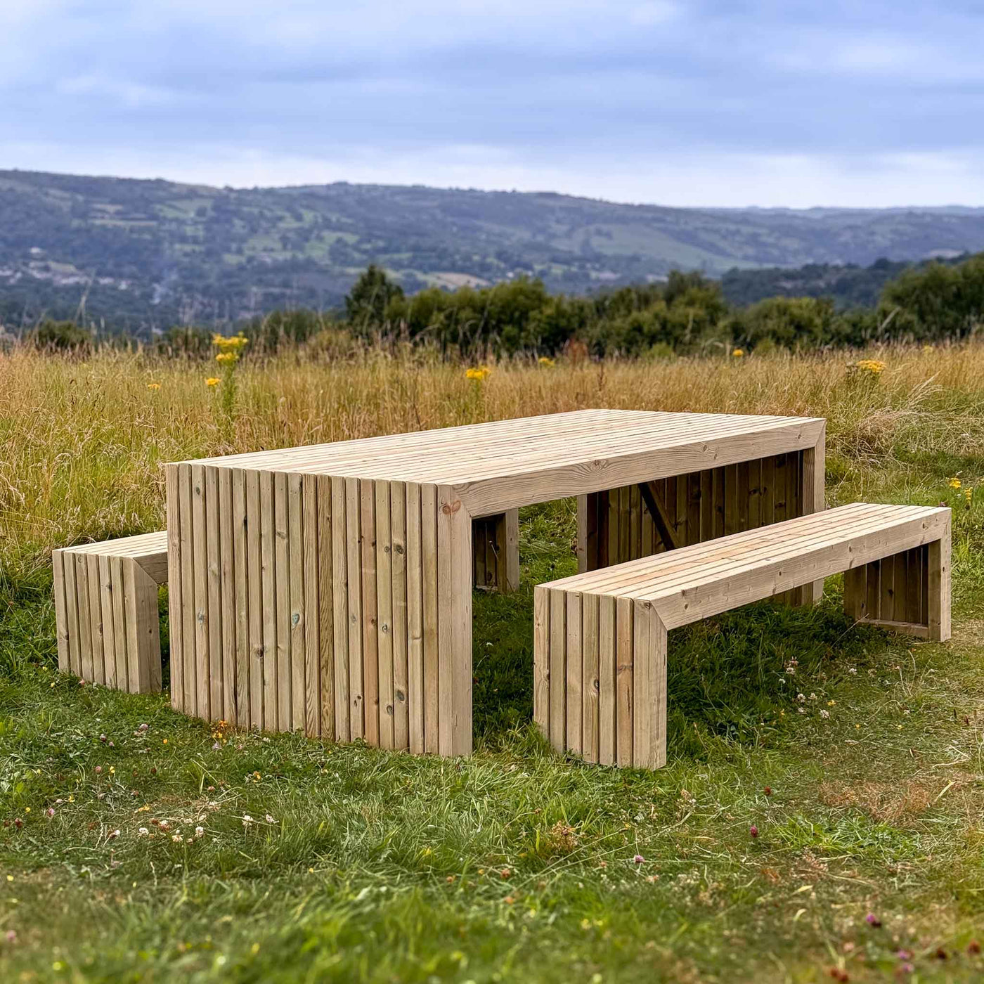 A durable, rustic wooden garden table set, perfect for outdoor dining and entertaining. The set features a long rectangular table with matching benches, all made from high-quality timber. The table and benches have a slatted design and are showcased in a scenic outdoor setting with rolling hills and a blue sky.