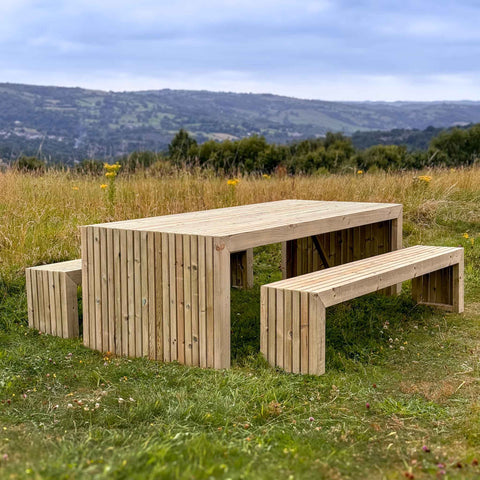 A durable, rustic wooden garden table set, perfect for outdoor dining and entertaining. The set features a long rectangular table with matching benches, all made from high-quality timber. The table and benches have a slatted design and are showcased in a scenic outdoor setting with rolling hills and a blue sky.