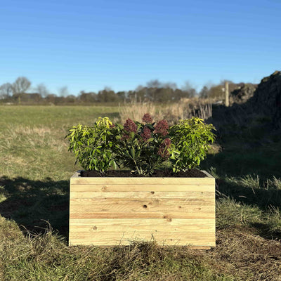 Front view of a rustic slatted wooden planter filled with green plants. Placed in a sunny garden by a stone wall.