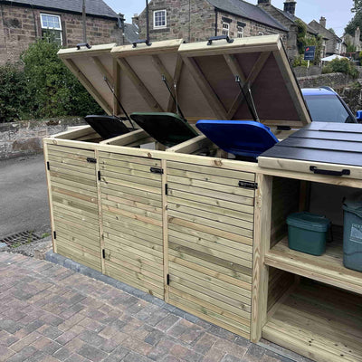 Wooden outdoor storage unit with doors open, containing recycling bins on a street.