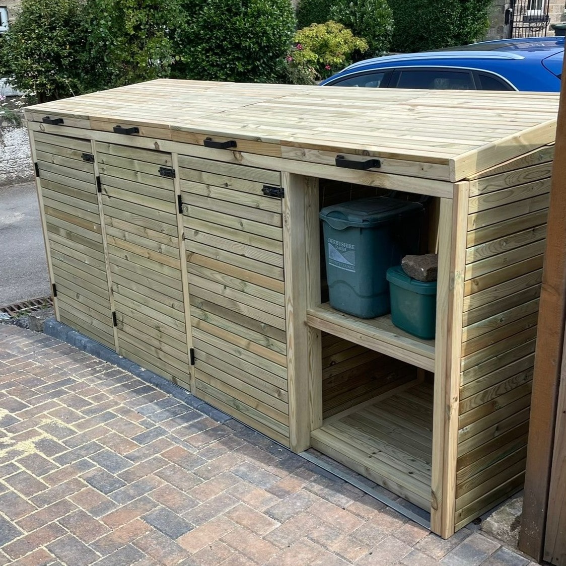 Wooden outdoor bin storage unit with doors open, showing green bins inside, on a paved driveway.