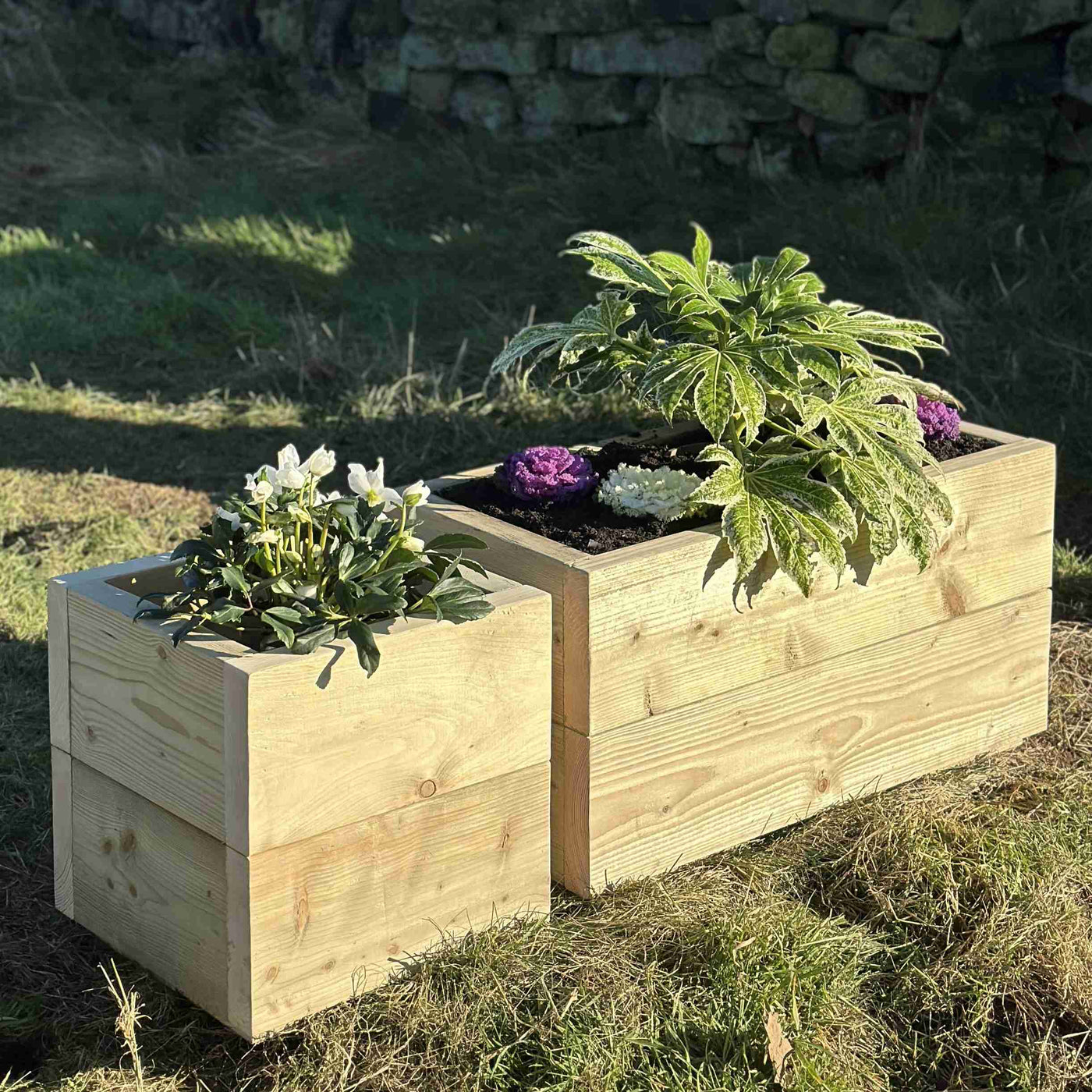 Heavy-duty wooden trough planters, one square and one rectangle filled with green plants and purple and white flowers. Placed on grass in front of a stone wall.