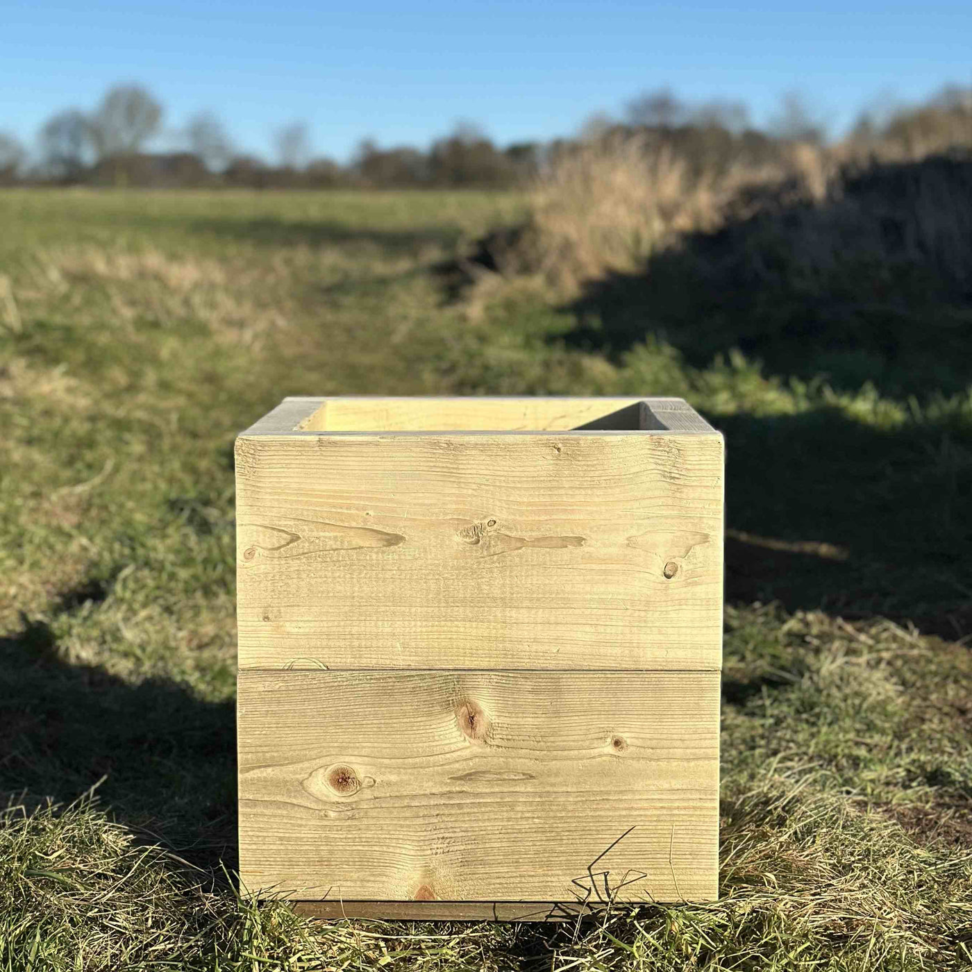 Square wooden trough planter, heavy-duty thick slatted design, no visable screws. A front view of the empty planter placed on grass.