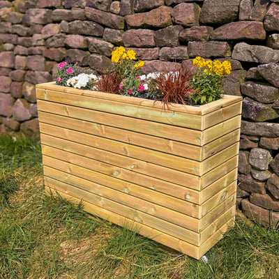 Corner view of a tall garden durable timber planter with bright flowers in front of a brick wall. 
