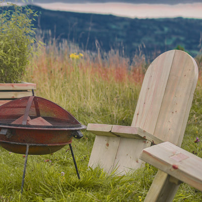 Wooden Adirondack Firepit Chairs surround a fire pit. The comfortable, chunky timber chairs are placed on lush green grass with a scenic backdrop of rolling hills.