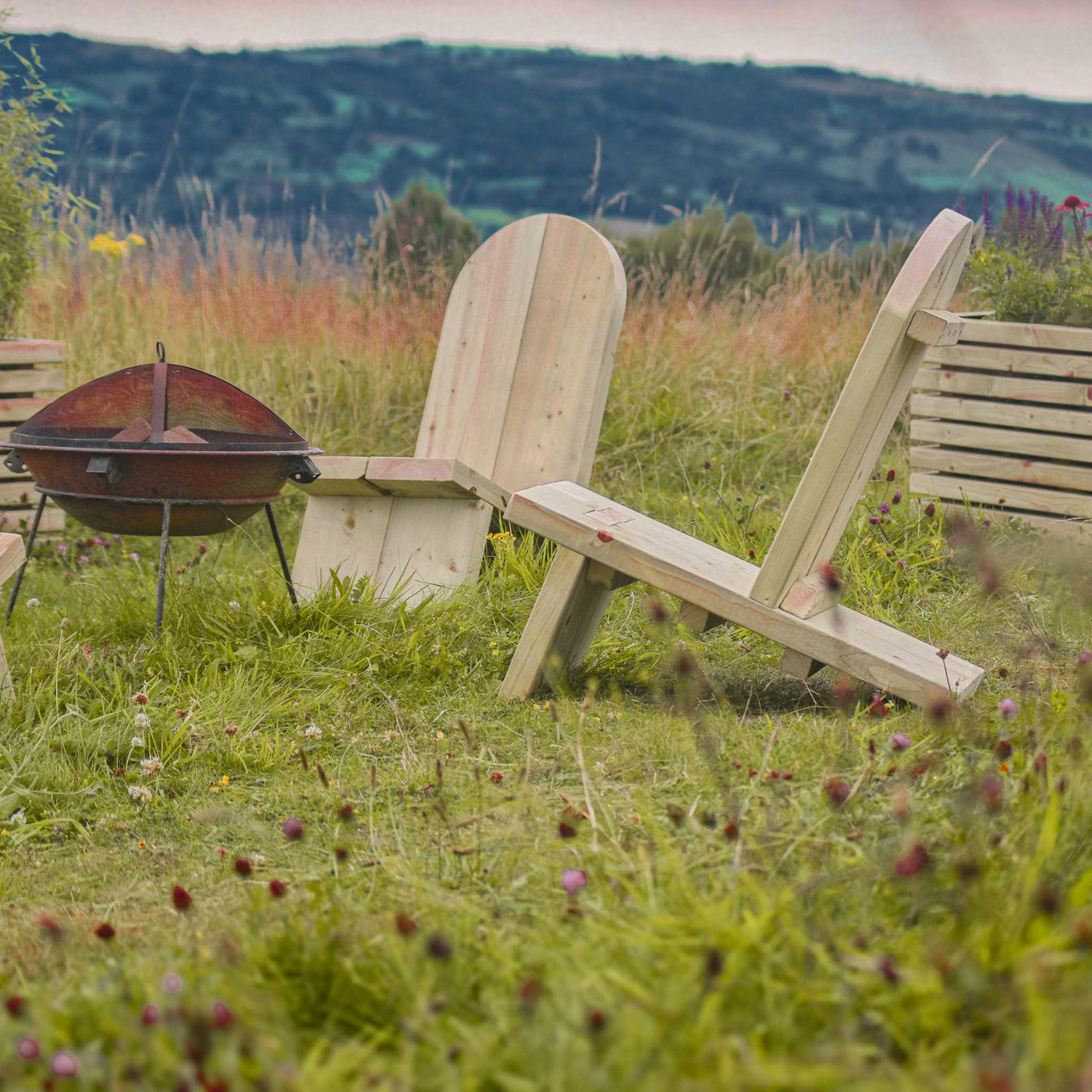 Wooden Adirondack Firepit Chairs surround a fire pit. The comfortable, chunky timber chairs are placed on lush green grass with two wooden planters behind them and rolling hills in the background.