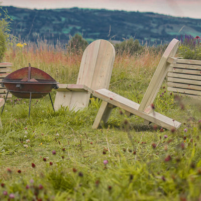 Wooden Adirondack Firepit Chairs surround a fire pit. The comfortable, chunky timber chairs are placed on lush green grass with two wooden planters behind them and rolling hills in the background.