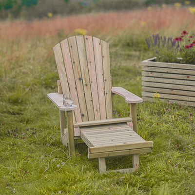 A wooden Adirondack Reclining Garden Chair with a foot extension is shown on a lush garden lawn to highlight its sustainable timber construction and outdoor comfort. A glass sits on the arm of the chair.