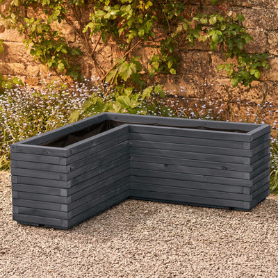 A wooden corner planter in an ironstone grey stain featuring an L-shaped design that fits perfectly into any space. Positioned in front of a rustic stone wall with green climbing branches, this planter is crafted from durable FSC-certified timber and comes pre-lined and ready for planting.