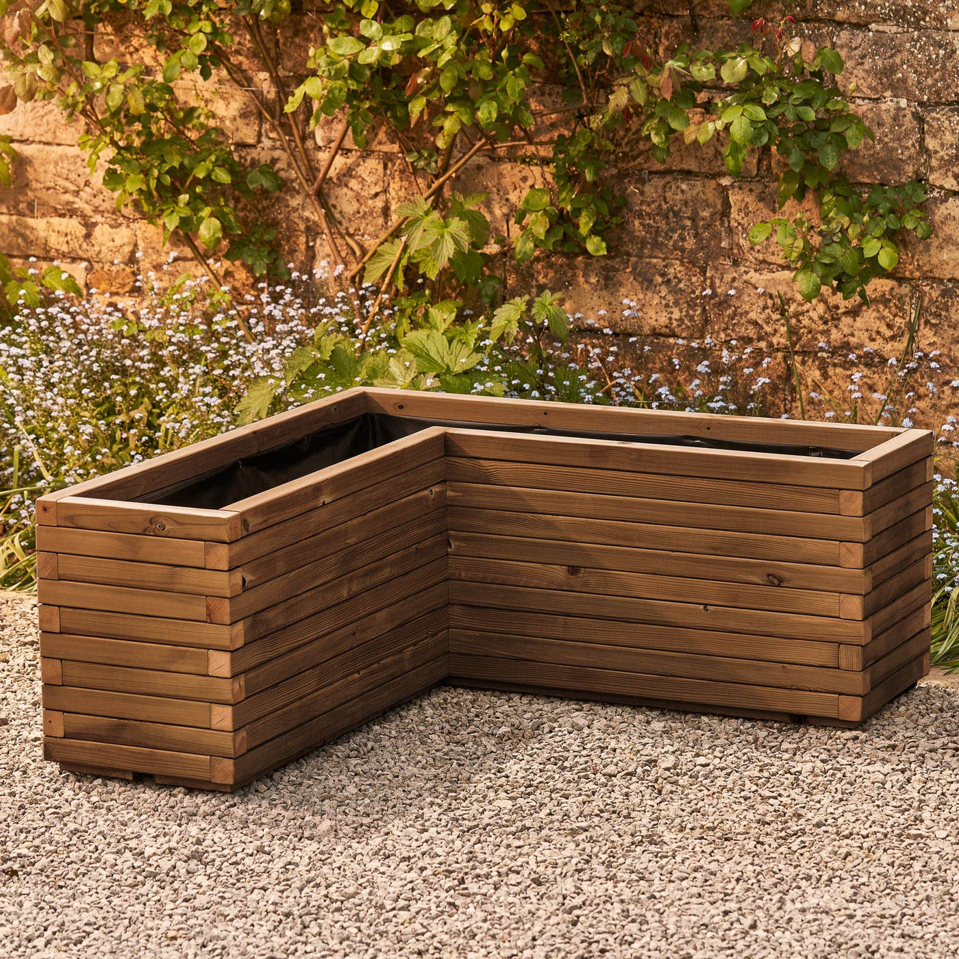 A wooden corner planter in a medium oak  stain featuring an L-shaped design that fits perfectly into any space. Positioned in front of a rustic stone wall with green climbing branches, this planter is crafted from durable FSC-certified timber and comes pre-lined and ready for planting.