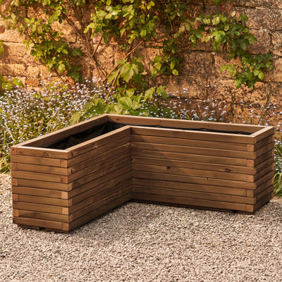 A wooden corner planter in a medium oak  stain featuring an L-shaped design that fits perfectly into any space. Positioned in front of a rustic stone wall with green climbing branches, this planter is crafted from durable FSC-certified timber and comes pre-lined and ready for planting.