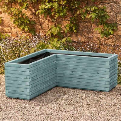 A wooden corner planter in a beach hut blue stain featuring an L-shaped design that fits perfectly into any space. Positioned in front of a rustic stone wall with green climbing branches, this planter is crafted from durable FSC-certified timber and comes pre-lined and ready for planting.