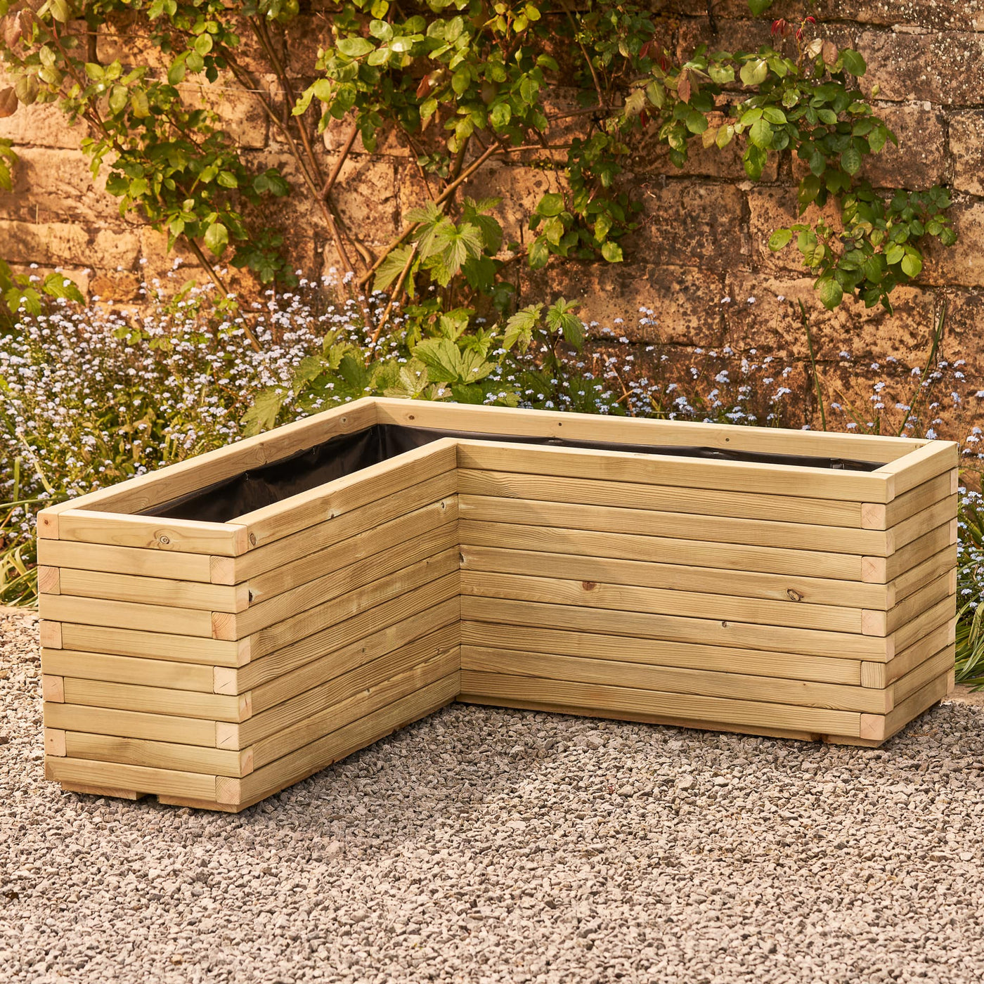 A wooden corner planter in a natural stain featuring an L-shaped design that fits perfectly into any space. Positioned in front of a rustic stone wall with green climbing branches, this planter is crafted from durable FSC-certified timber and comes pre-lined and ready for planting.