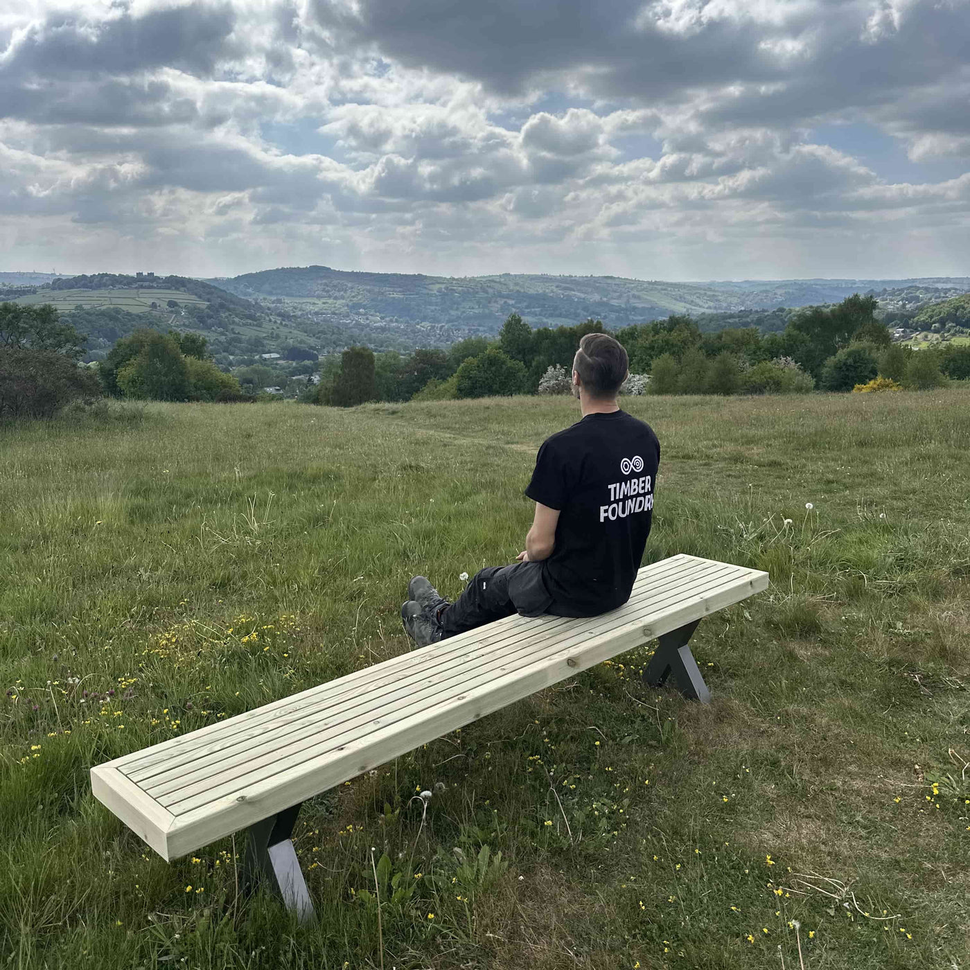 Man sitting on a wooden garden bench with steel x legs with rolling hills in the background, part of the Wooden Garden Dining Set.